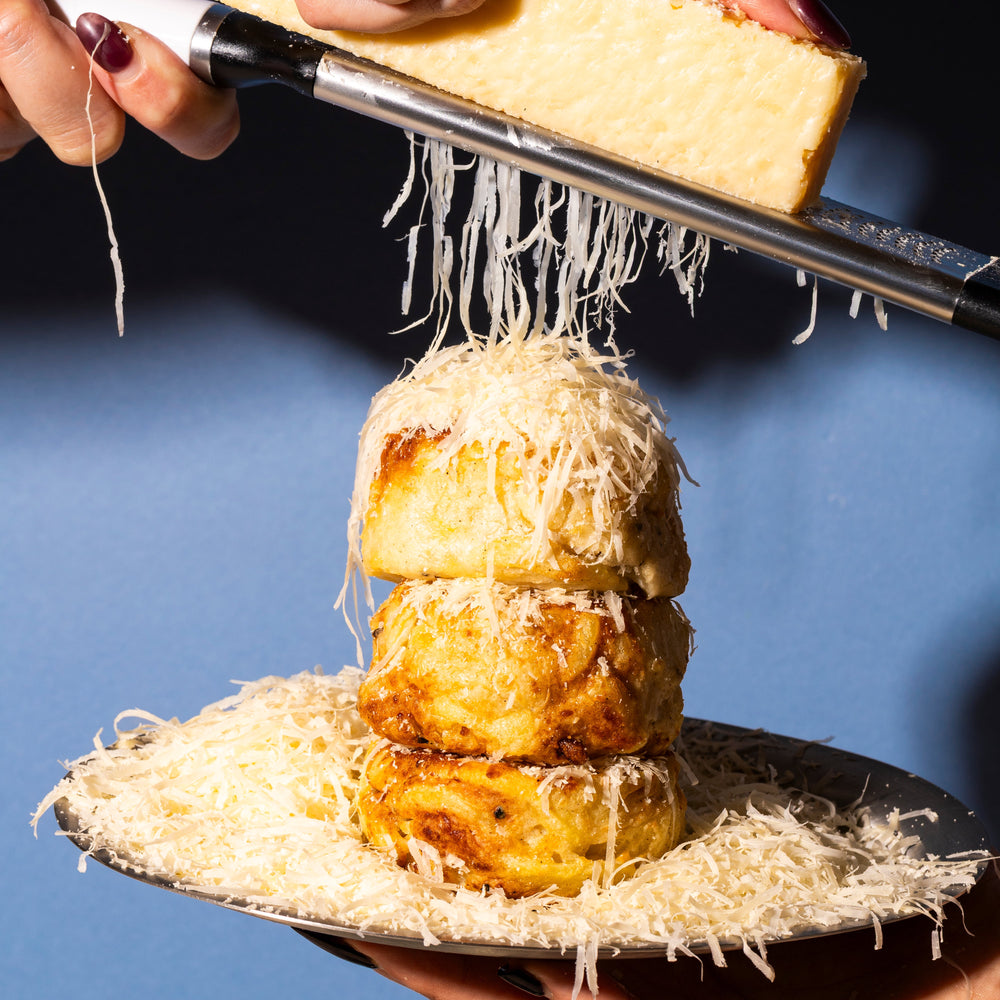 Person grating Parmesan cheese onto a stack of Potato Utopia Gratins on a blue background