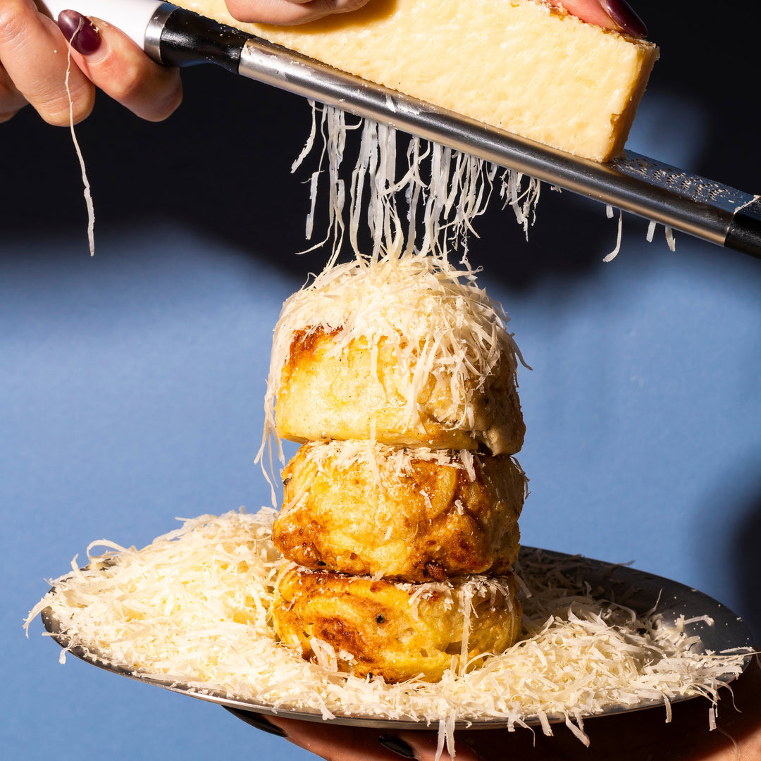 Person grating Parmesan cheese onto a stack of Potato Utopia Gratins on a blue background